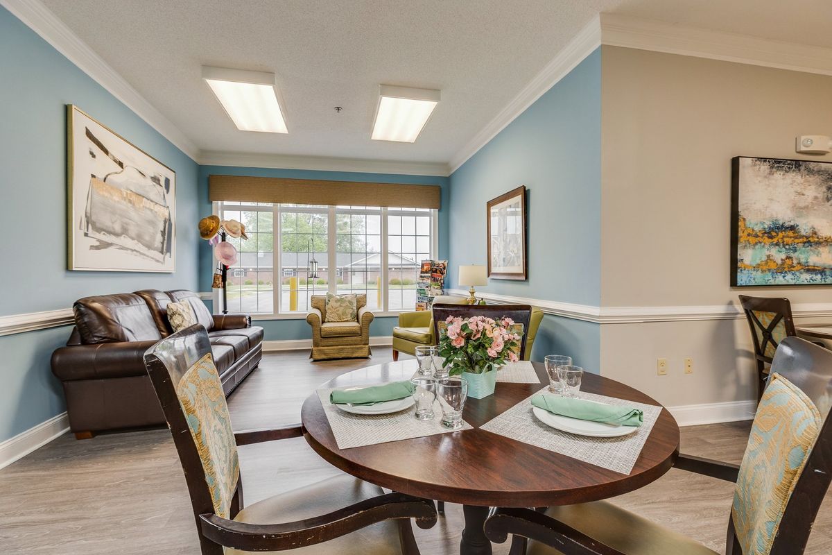 A cozy living and dining area with blue walls, featuring a set dining table, sofa, armchair, and large windows allowing natural light.