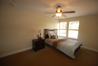 A cozy bedroom with a bed, nightstand, and ceiling fan, illuminated by natural light from the windows.