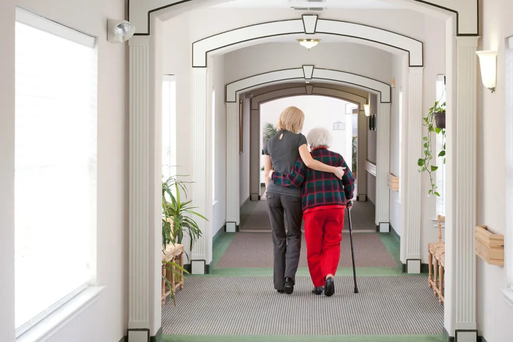 A caregiver assists an elderly person walking down a hallway in a nursing facility.