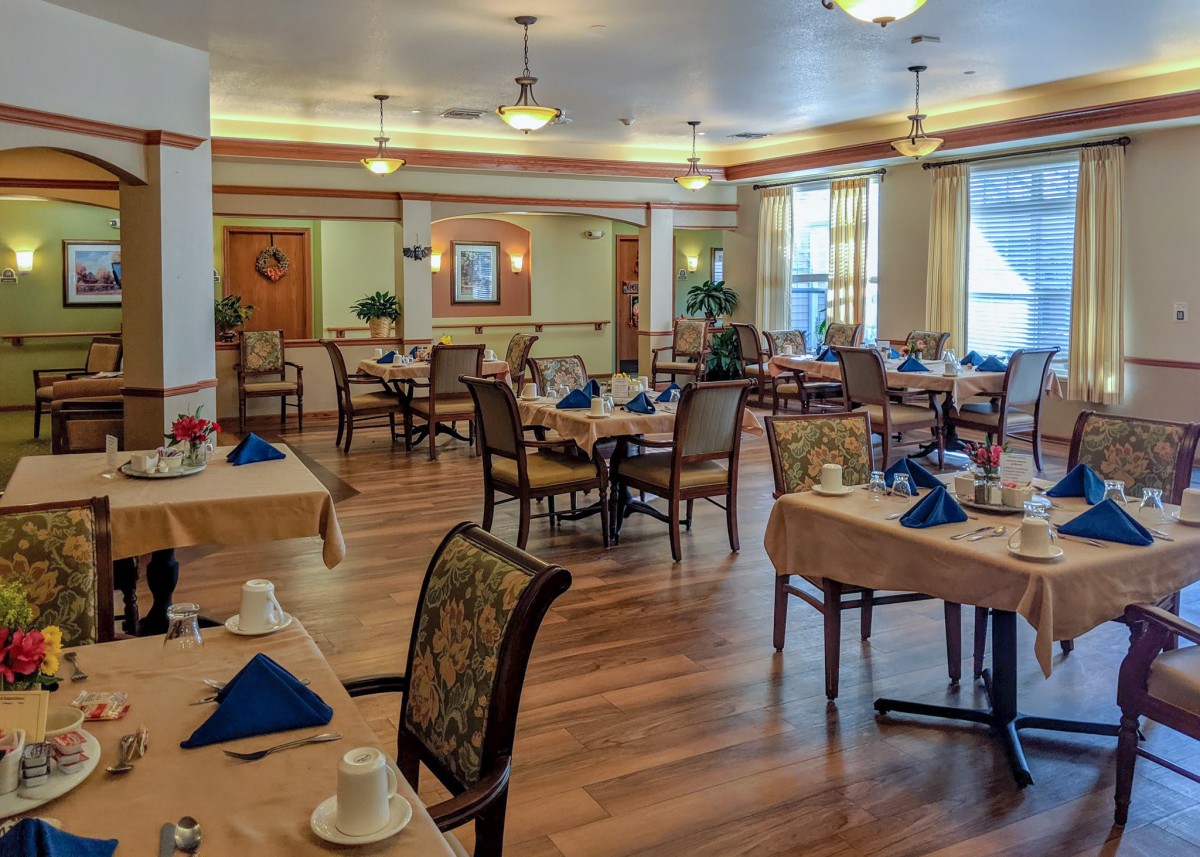 A well-lit dining room with neatly arranged tables and chairs, featuring elegant table settings on a wooden floor.
