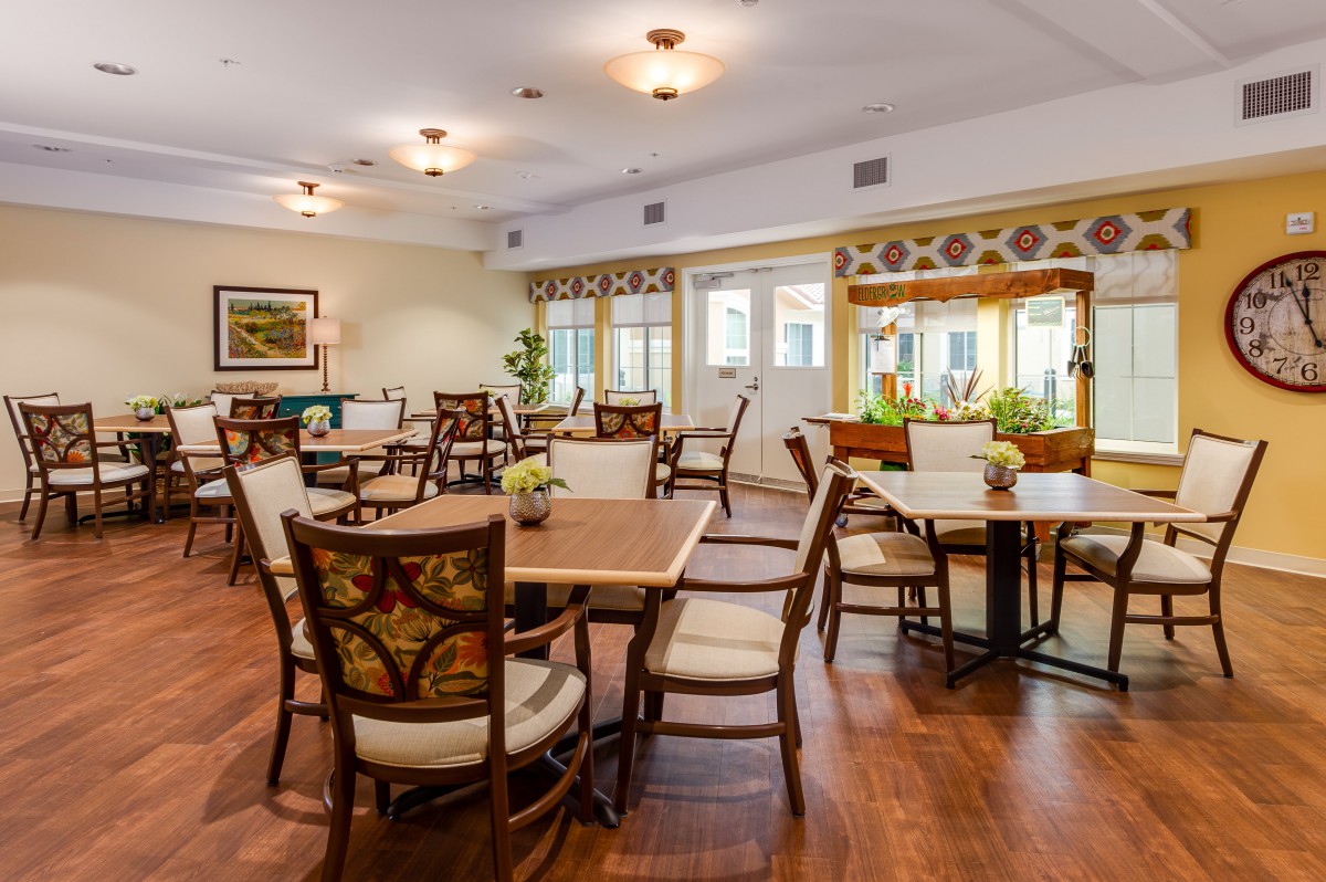 A well-lit dining room with several tables and chairs arranged on a wooden floor, featuring decorative elements and large windows.