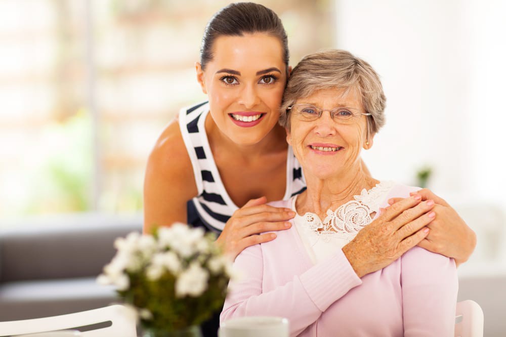 A smiling younger woman embraces an elderly woman indoors, showcasing a warm family bond.