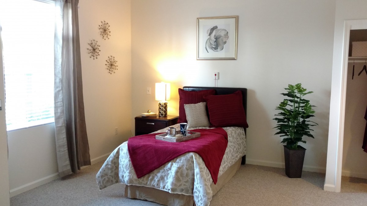 A cozy bedroom with a single bed, featuring red and beige decor, a lamp on the nightstand, and wall art.