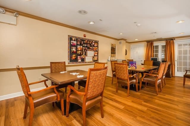 A cozy community dining area with wooden floors, tables, and chairs, featuring a bulletin board on the wall.