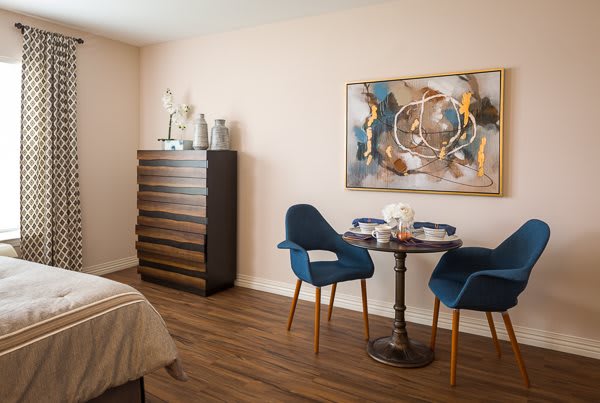 A modern bedroom featuring a cozy dining area with two blue chairs, a small round table, and abstract artwork on the wall.