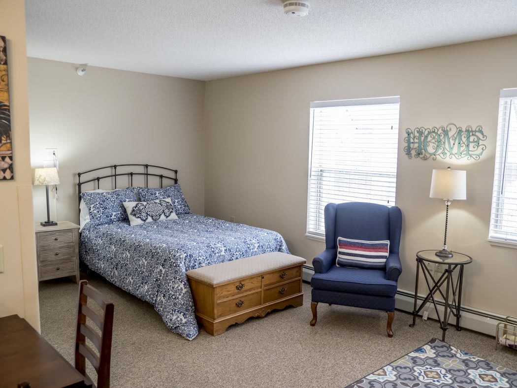A cozy bedroom with a metal bed frame, patterned bedding, an armchair, and a window allowing natural light.
