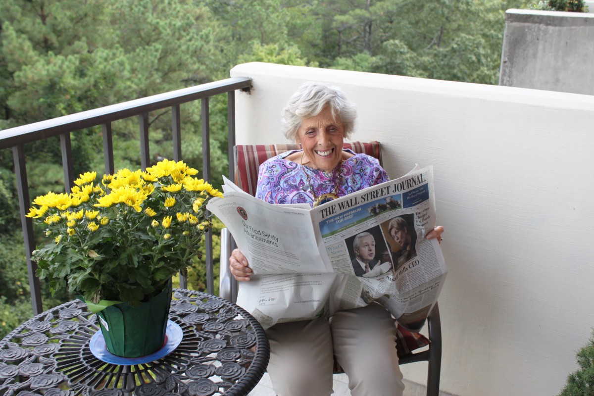 An elderly woman relaxes on a balcony while reading a newspaper, surrounded by flowers.