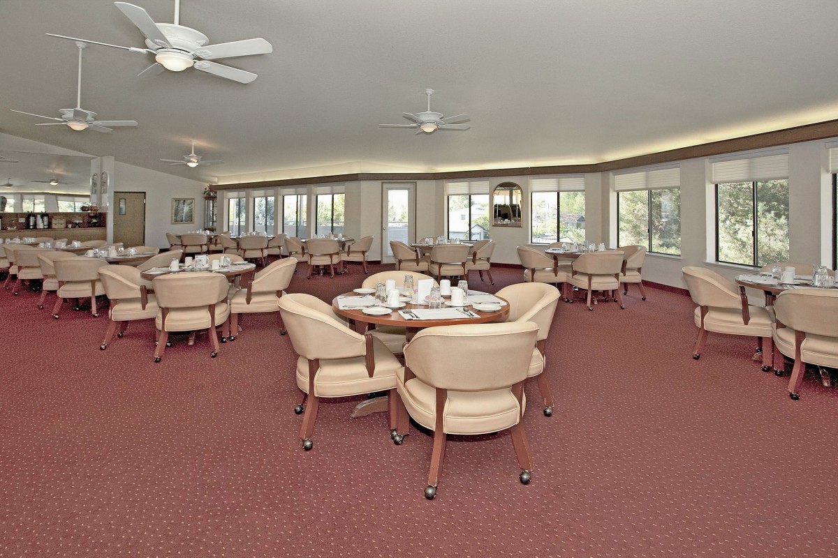 A spacious dining room with multiple tables and chairs, featuring large windows and ceiling fans.