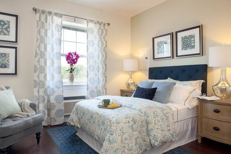 A cozy bedroom with elegant decor, featuring a bed with patterned bedding, a lamp on the nightstand, and a window adorned with curtains and flowers.
