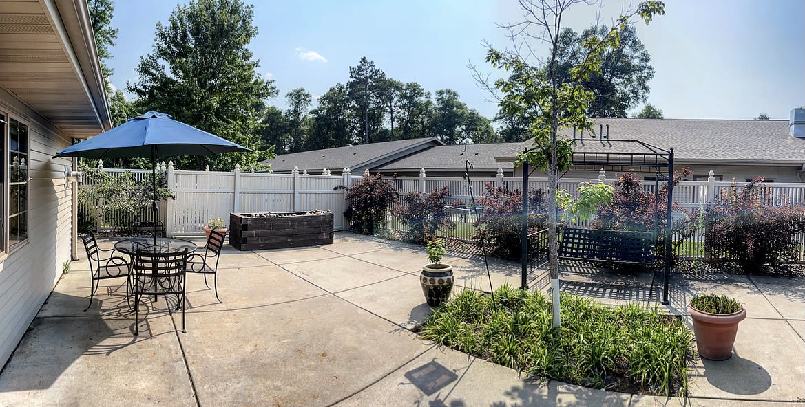 A sunny backyard patio featuring outdoor furniture, a garden with potted plants, and a surrounding fence.