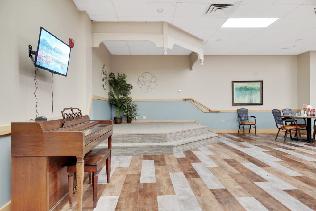A spacious room with a piano, stage, and seating area, featuring wooden flooring and decorative plants.