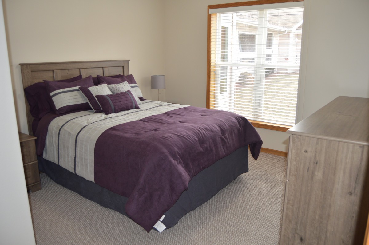 A cozy bedroom with a neatly made bed, featuring purple and gray bedding, a bedside lamp, and a large window with blinds.
