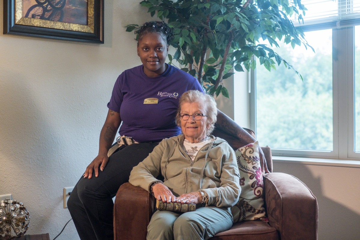 A caregiver and an elderly person sitting together indoors, smiling for the photo.