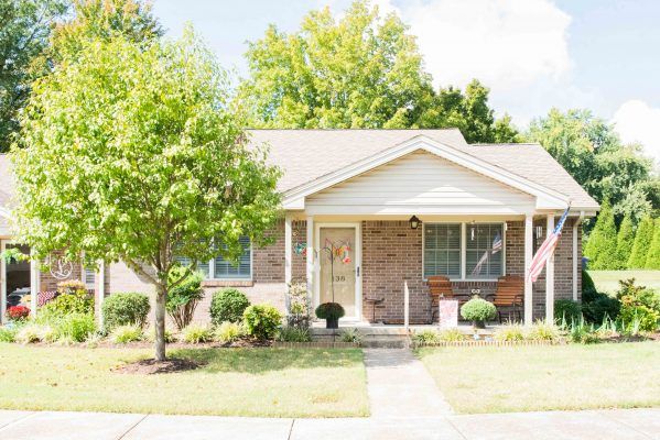 A charming brick house with a small porch, surrounded by trees and a well-maintained lawn.