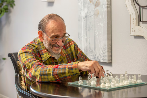 An elderly man smiles as he thoughtfully moves a piece on a glass chess set.