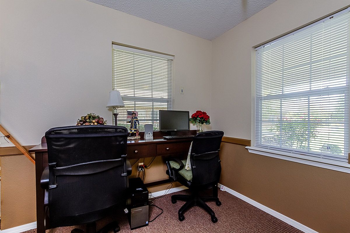 A small office workspace with a desk, computer, and chairs positioned by windows.
