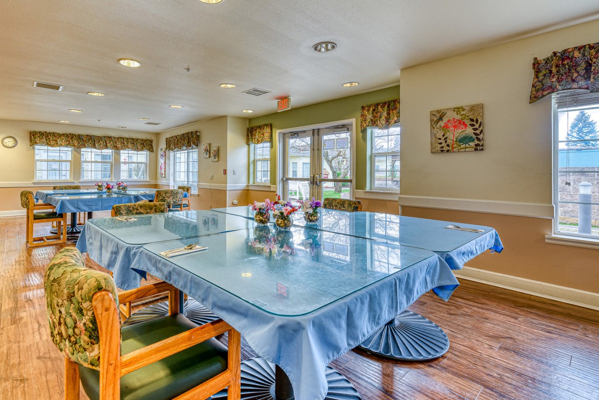 A well-lit dining area with tables and chairs, accented by natural light from large windows and simple decor.