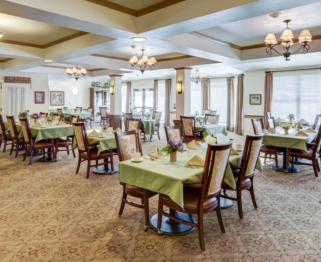 Spacious dining room with elegantly set tables and chairs, featuring chandeliers and large windows.