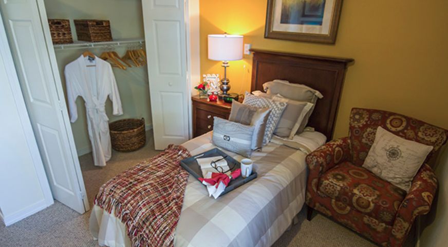 A cozy bedroom featuring a neatly made single bed with a tray of breakfast, a table lamp on the nightstand, and a robe hanging in the open closet.