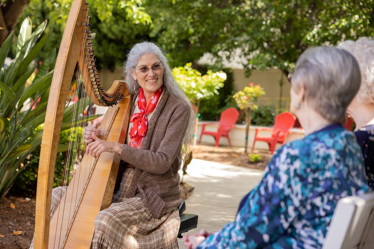 An elderly woman plays the harp in a garden setting, entertaining another person.