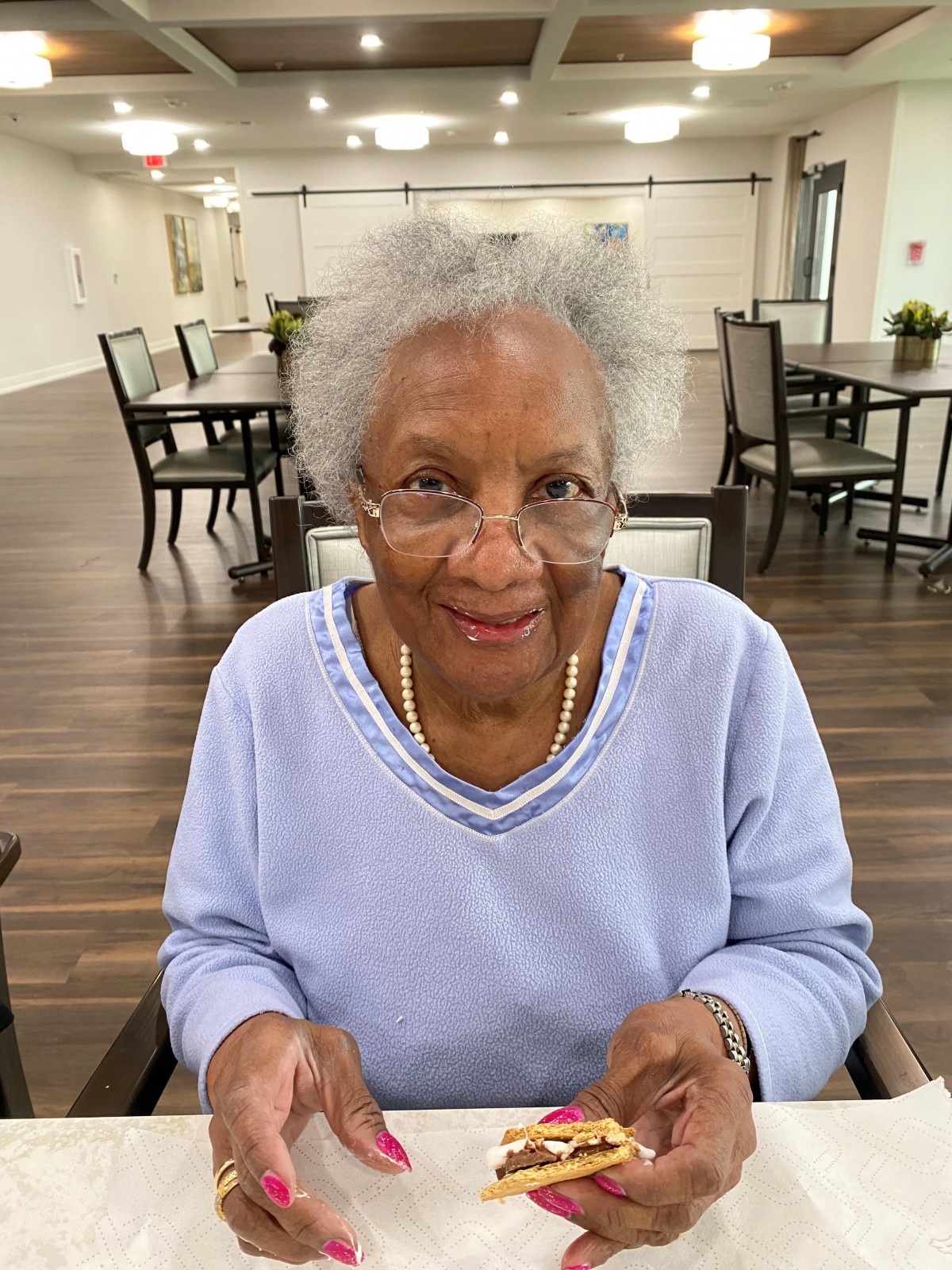 An elderly woman with gray hair and glasses, smiling in a dining area.