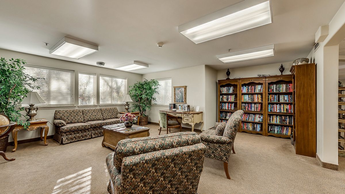 A cozy waiting room with patterned sofas, a wooden table, a bookshelf filled with books, and a potted plant.
