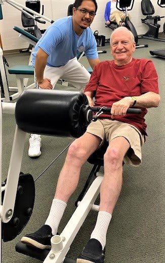 An elderly man smiling while exercising on a machine at the gym.