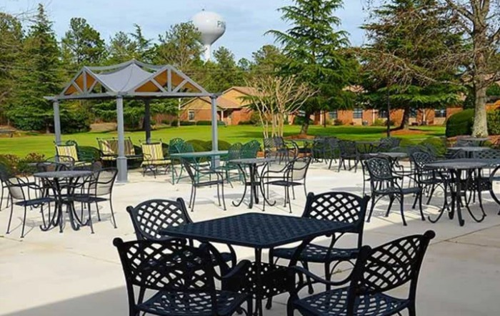 An outdoor seating area with metal tables and chairs, set in a park-like environment with greenery and trees.