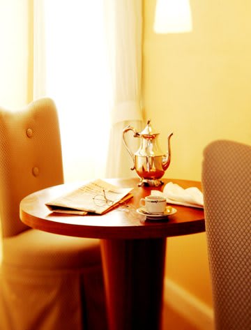 A cozy setting with a teapot and cup on a round wooden table, surrounded by upholstered chairs and warm lighting.