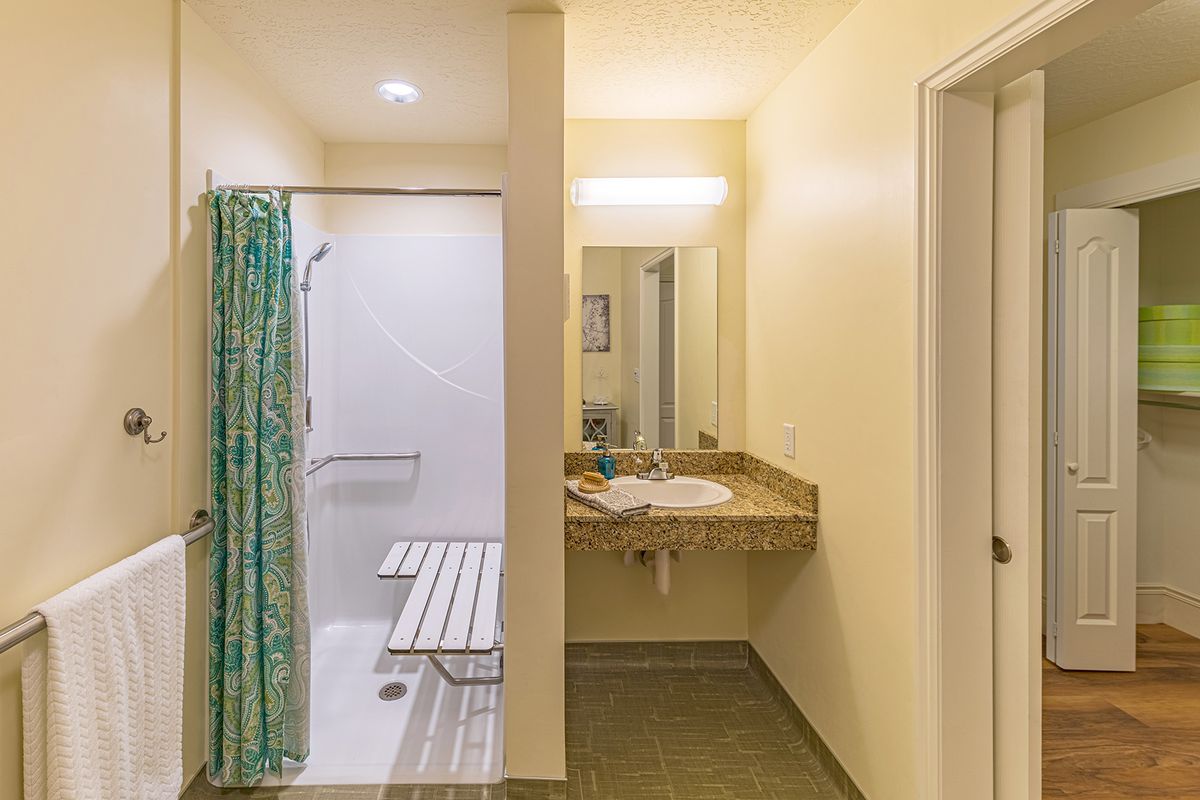 An accessible bathroom with a shower featuring safety bars and a bench, adjacent to a sink and mirror.