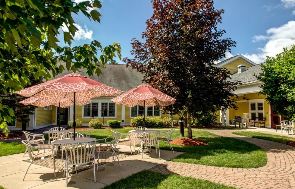 A charming outdoor patio area with tables and chairs shaded by red umbrellas, surrounded by trees and a yellow building.