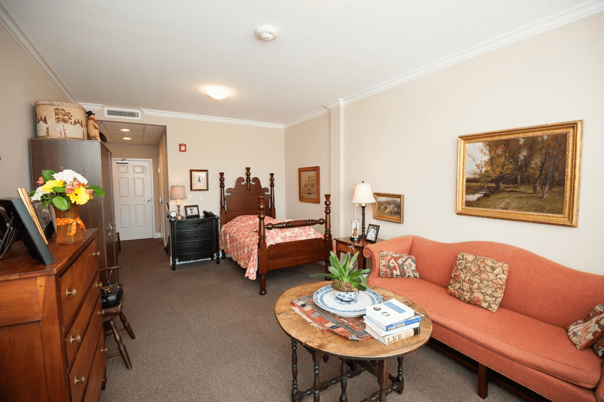 A cozy studio apartment with a combined living and sleeping area, featuring a sofa, round table, bed, and decorative elements like artworks and plants.
