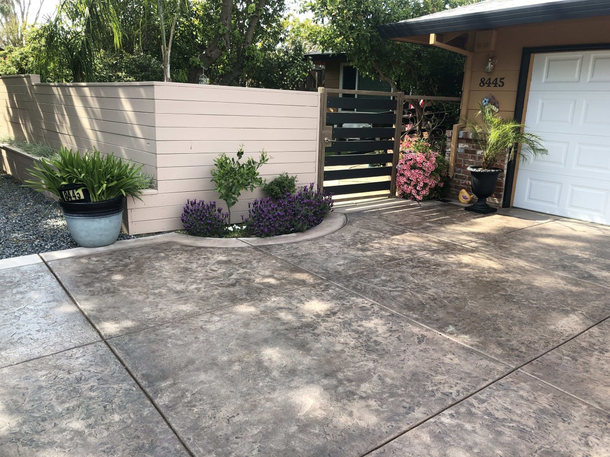 A clean concrete driveway with potted plants and a wooden fence, leading to the entrance of a house.