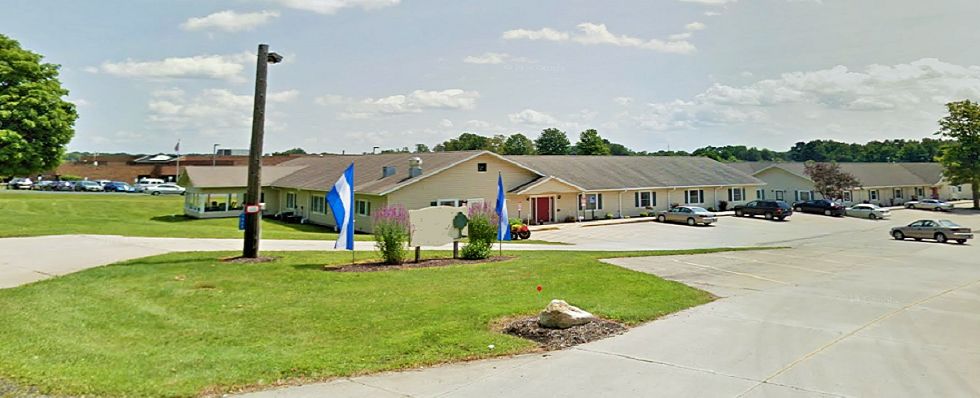 A single-story building with two blue and white flags near the entrance, surrounded by greenery and a clear sky above.