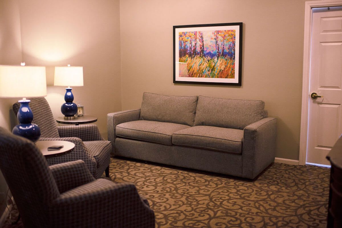 A cozy living room featuring a gray sofa, an armchair, a table lamp, and a colorful painting on the wall.