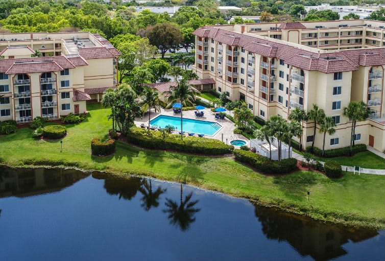 Aerial view of a residential apartment complex with a pool, surrounded by lush greenery and a tranquil lake.