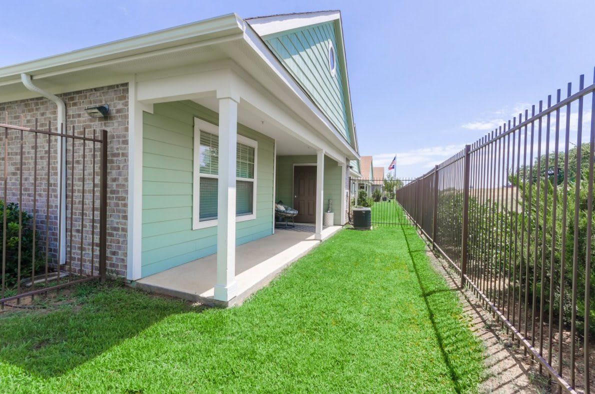 A small house with a well-maintained lawn and porch, bordered by a fence under a clear blue sky.