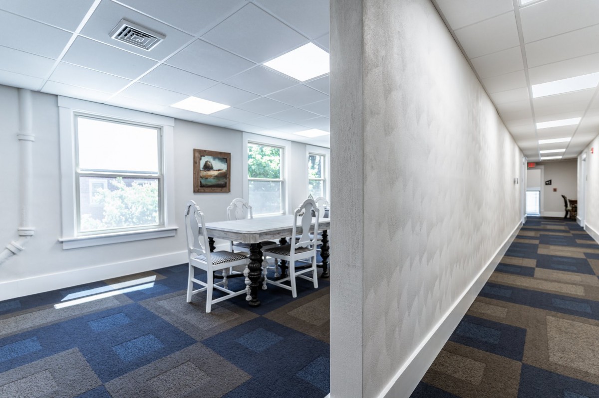 A bright meeting room with white chairs and a table, featuring large windows and artwork on the wall.