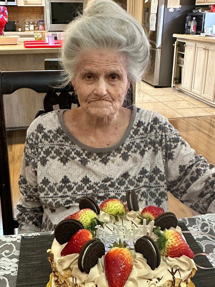 An elderly woman sitting at a table with a decorated birthday cake in front of her.