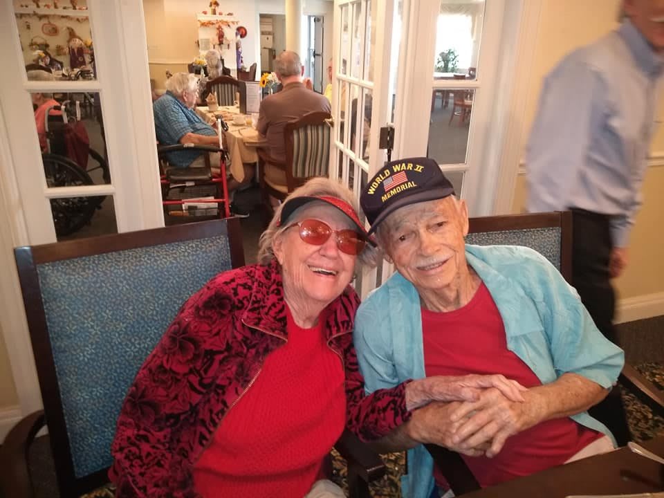 An elderly couple smiling and holding hands in a cozy indoor setting.