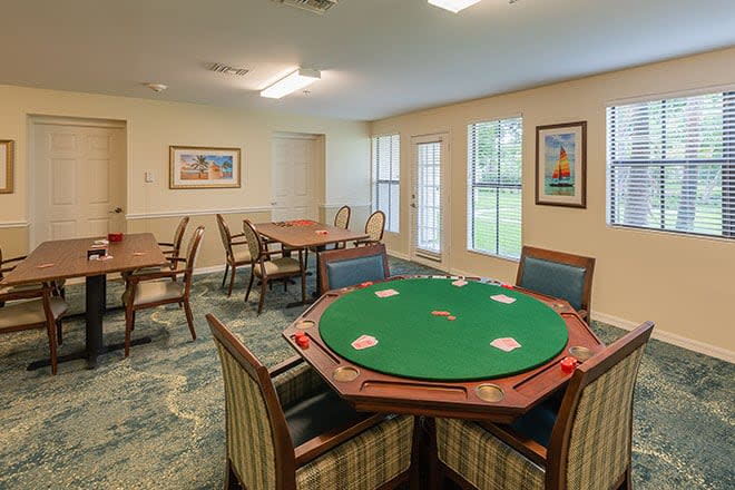 A cozy game room featuring a poker table surrounded by chairs, with large windows and artwork on the walls.