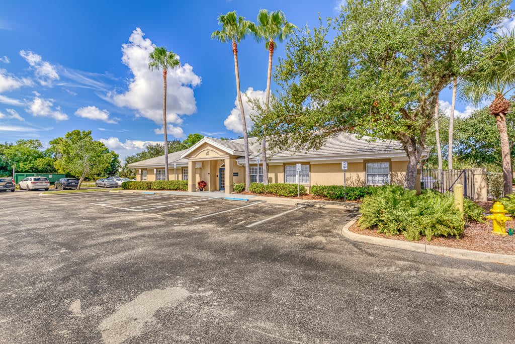 A single-story building with a parking lot surrounded by palm trees and greenery under a bright blue sky with clouds.