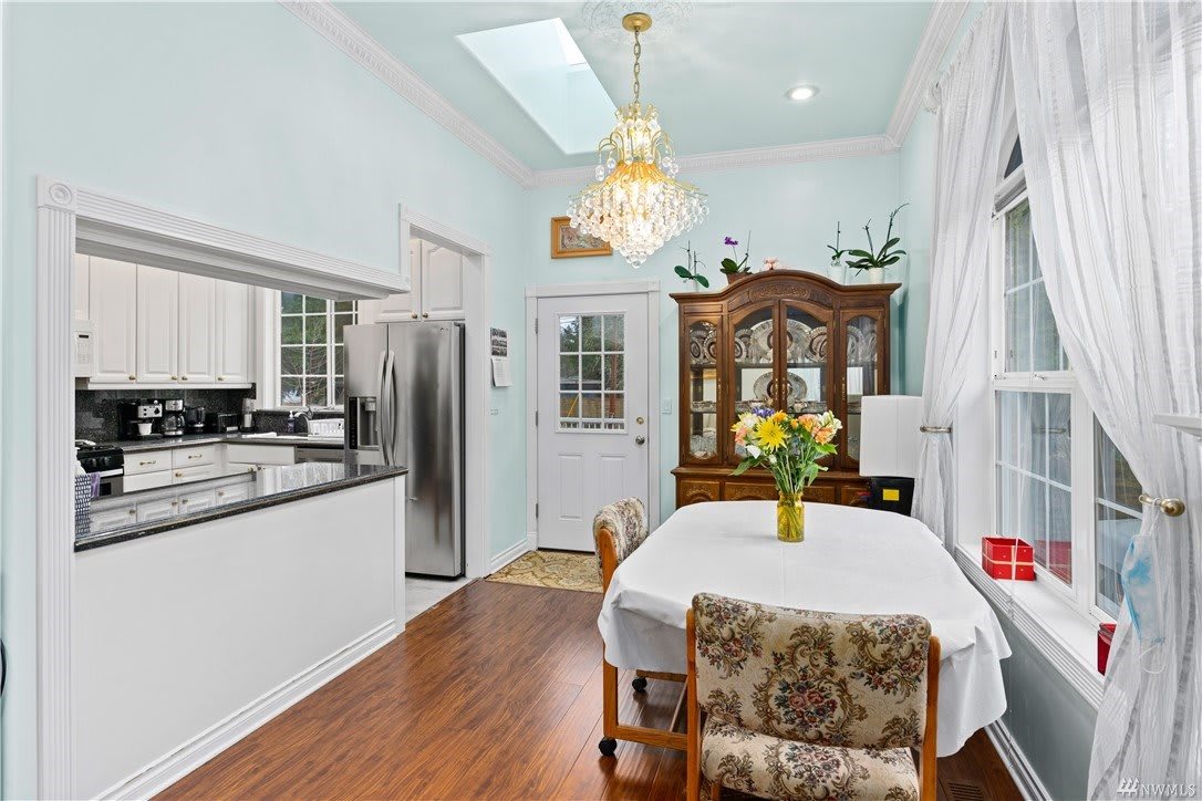 A cozy kitchen and dining area featuring a table set with chairs, an elegant chandelier, and a wooden cabinet, all framed by soft white walls and a skylight.
