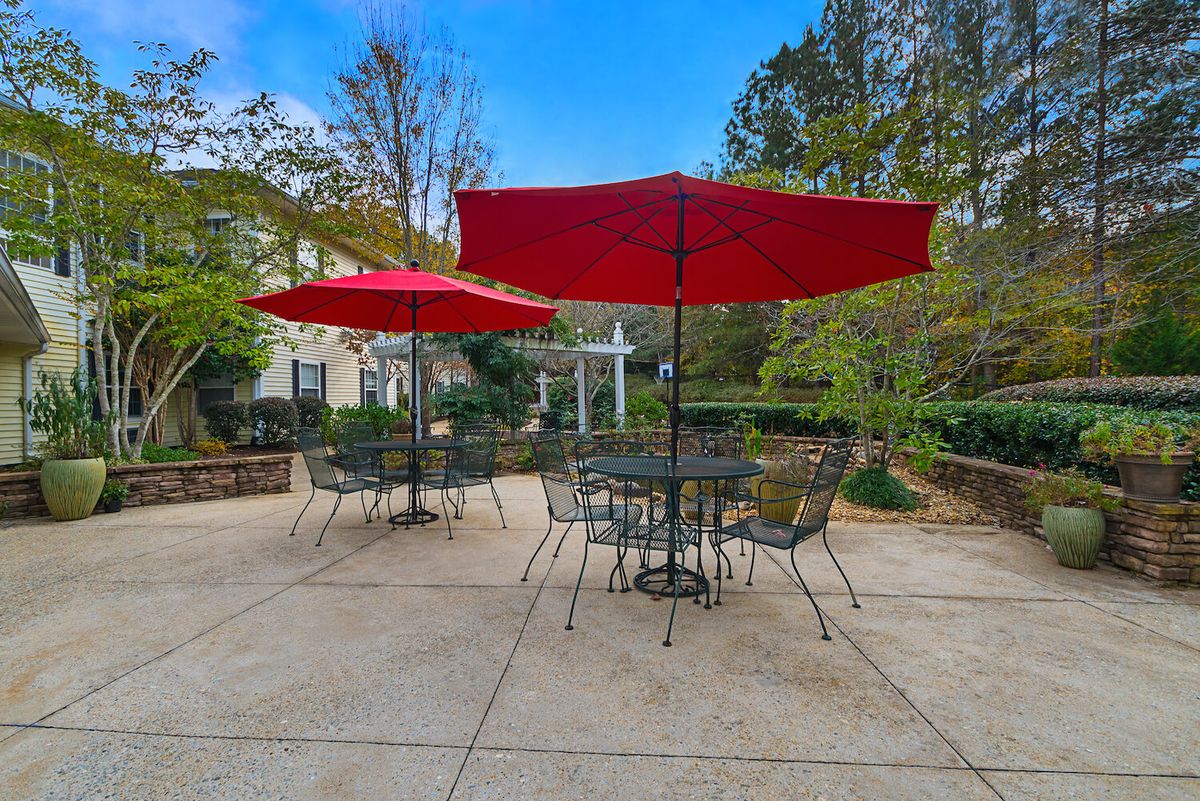 Outdoor patio area with tables, chairs, and red umbrellas surrounded by greenery.