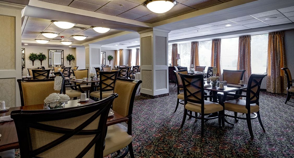 An elegant dining area with multiple tables and chairs, featuring carpeted flooring and ambient lighting.