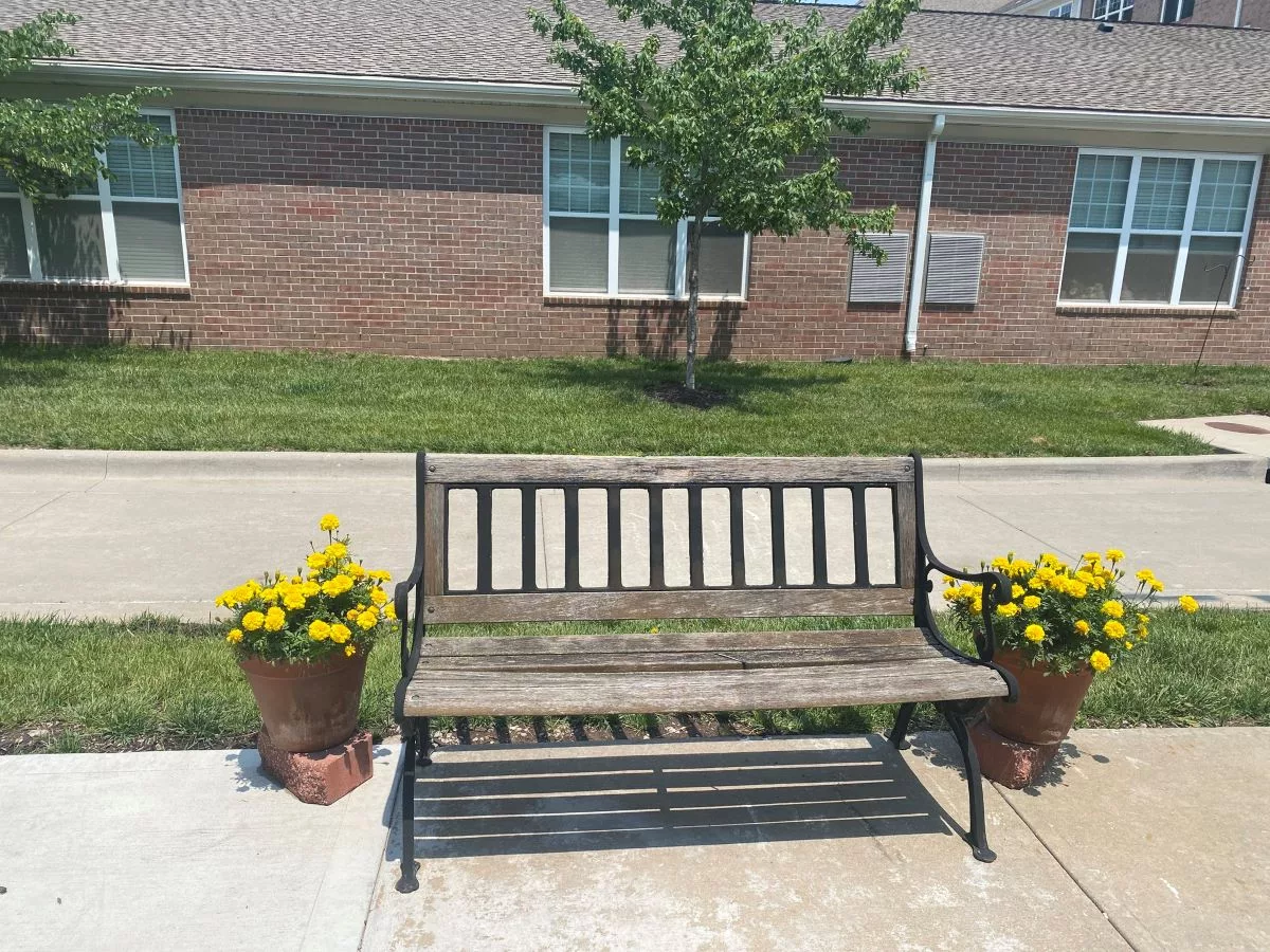 A wooden bench flanked by potted yellow flowers on a sunny day, set against a brick building.