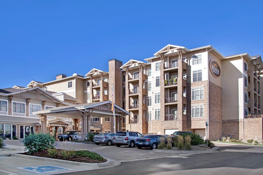 A modern multi-story apartment building with a parking area in front, set against a clear blue sky.