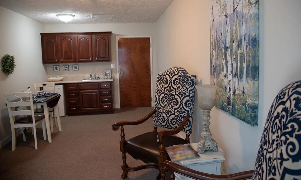 A cozy living space featuring a patterned chair, artwork on the wall, and a kitchenette in the background.
