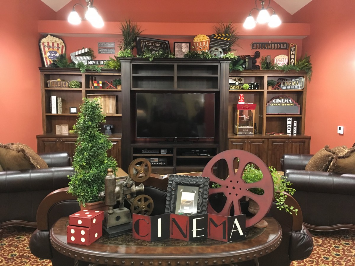 A cozy home theater setup with cinema-themed decor, featuring a vintage film reel, bookshelves, and greenery around a television.