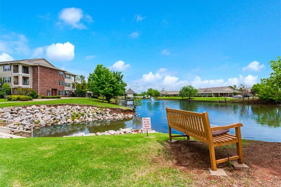 A serene lakeside view with a wooden bench, surrounded by houses and lush greenery under a bright blue sky.